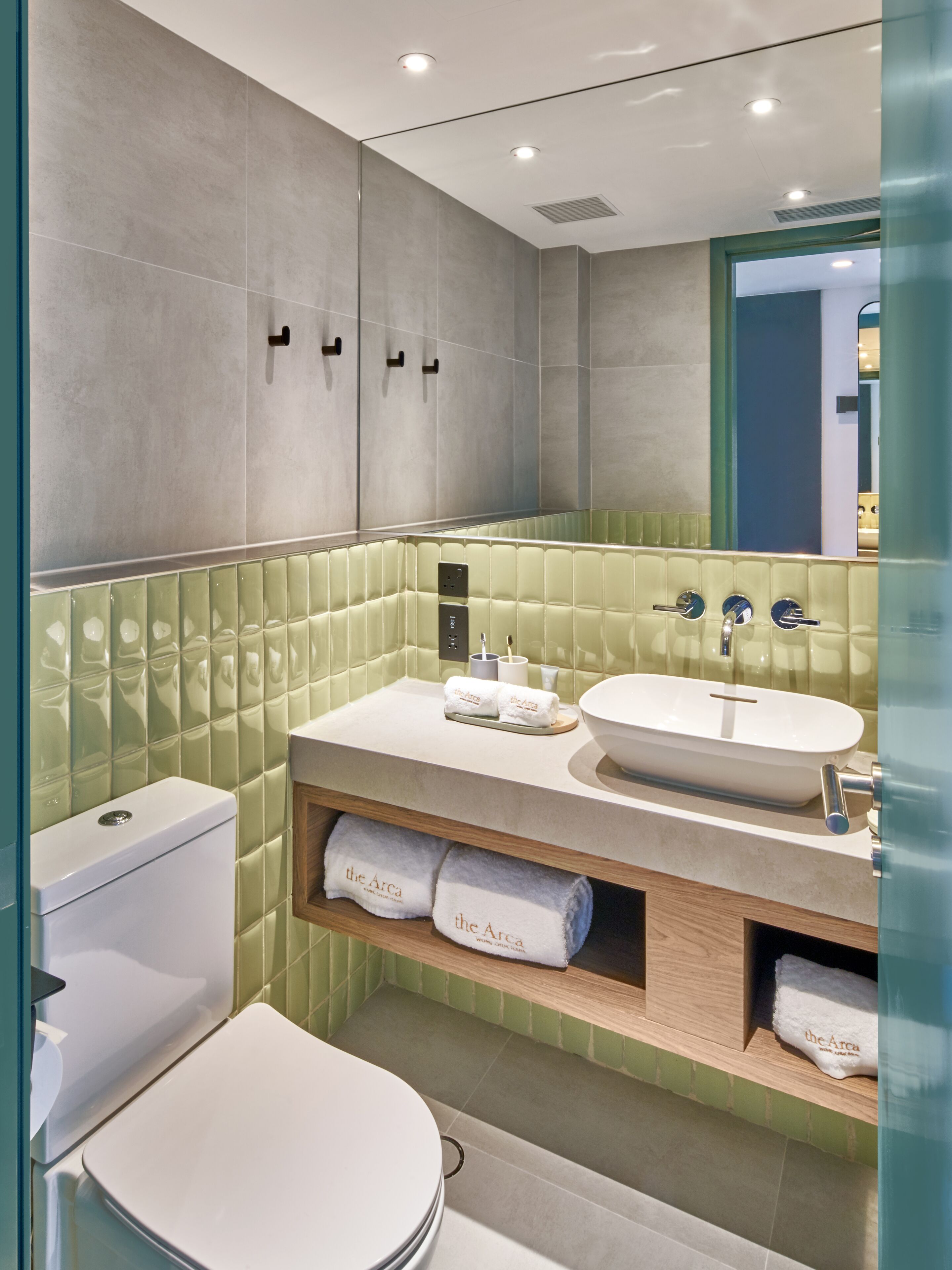 A modern bathroom with a white toilet, a beige sink with a large basin, and a green-tiled wall. There are two towel racks with towels, and a mirror above the sink.