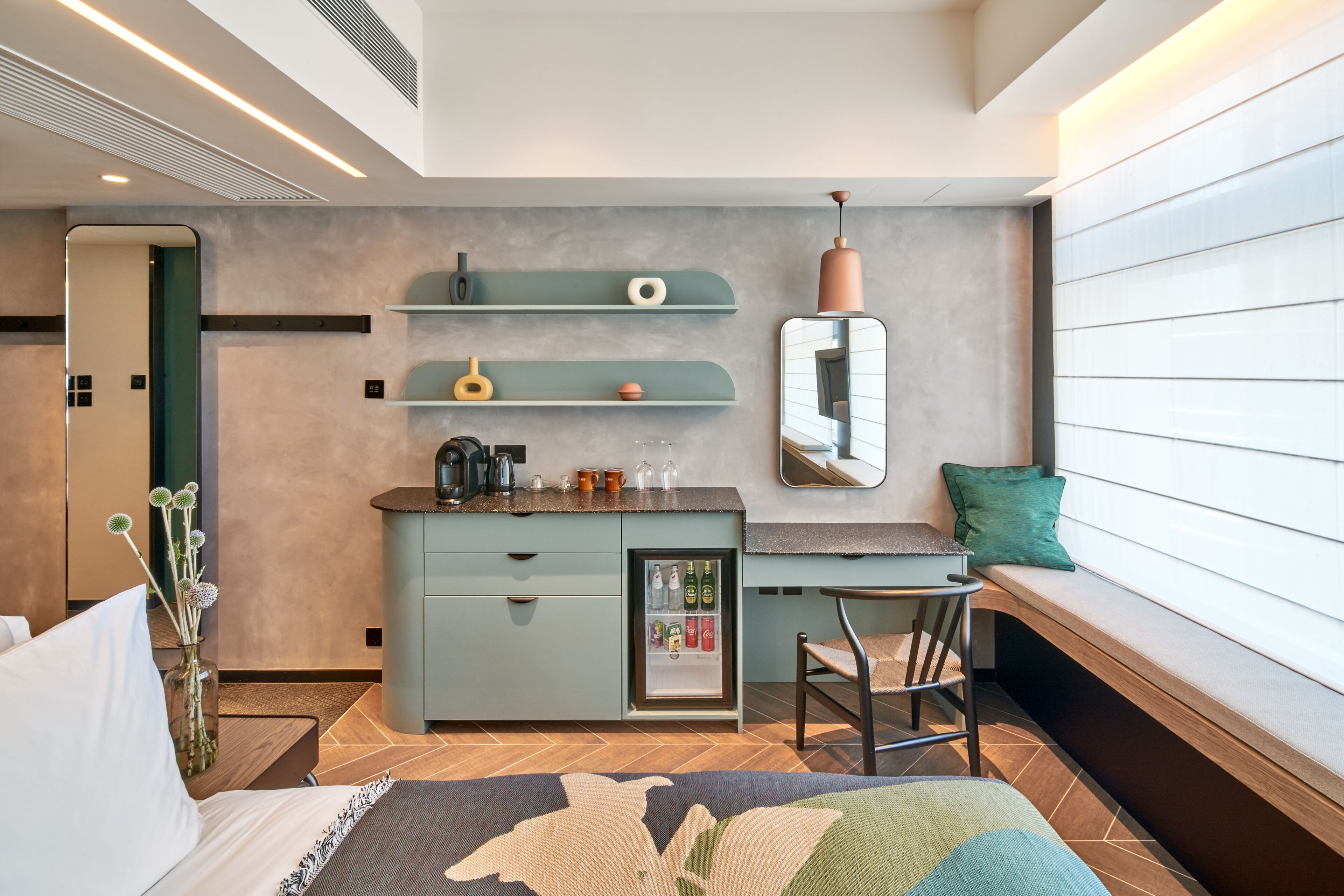 A modern hotel room with a kitchenette, a seating area with a bench, and a small refrigerator stocked with beverages.
