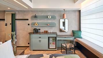 A modern hotel room with a kitchenette, a seating area with a bench, and a small refrigerator stocked with beverages.