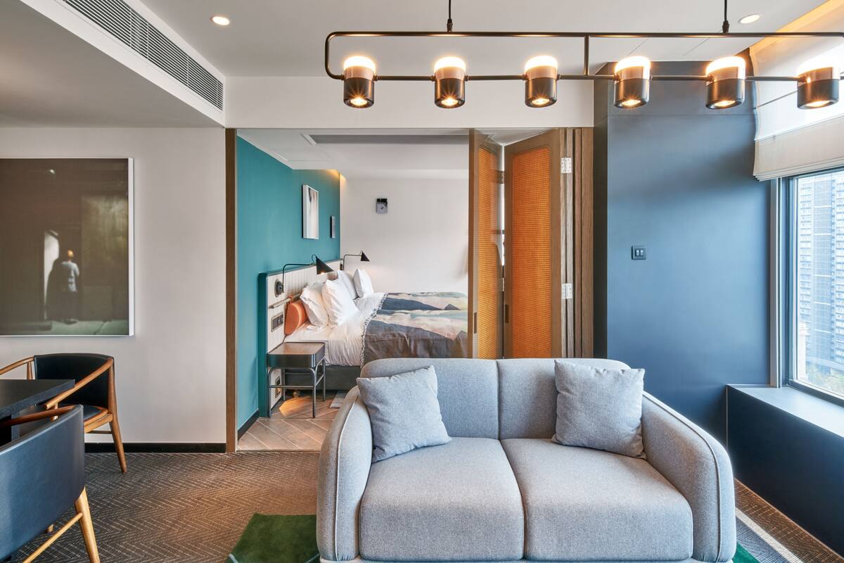 Una habitación de hotel moderna con un sofá gris, una mesa de comedor y un ventanal grande.