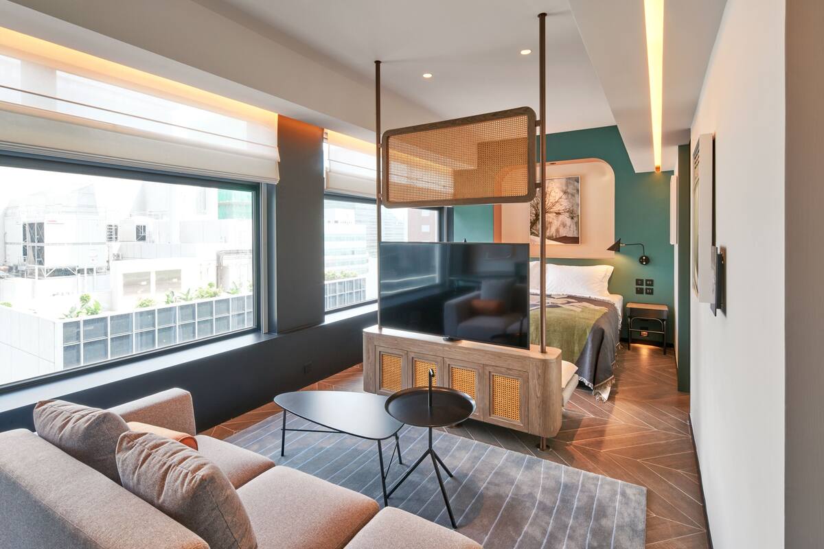 Una habitación de hotel moderna con un ventanal grande, un sofá, una mesita, un televisor y una cama.