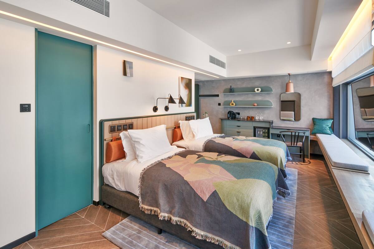 Una habitación de hotel moderna con dos camas, una kitchenette, un rincón de descanso junto a la ventana y una puerta color turquesa.
