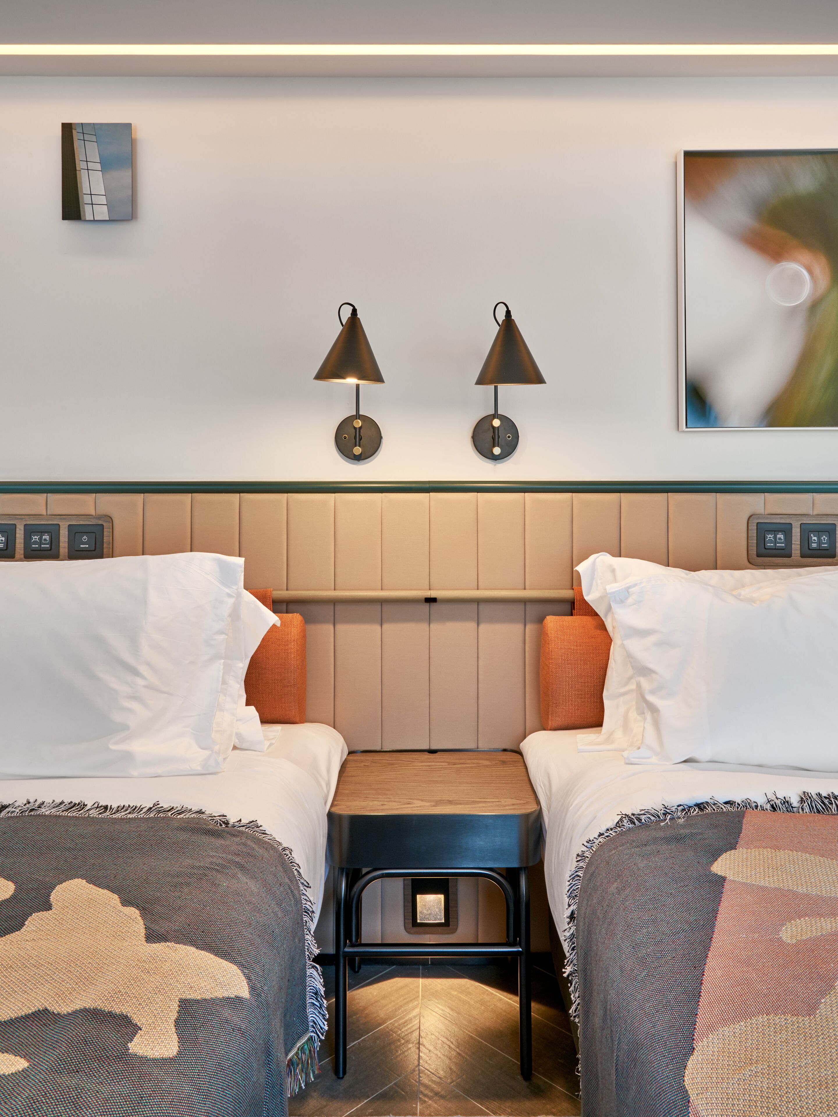 A hotel room with two beds, a small wooden table, and two wall-mounted lamps.