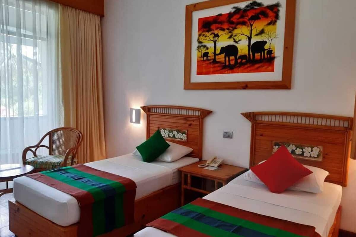 Habitación de hotel con dos camas individuales, cabecera de madera, mesita de noche con teléfono, una silla y una ventana con cortinas.