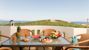 A table with breakfast items, wicker chairs, and a view of a green hill and sea. - Panoramic Sea Views, Pool, Garden, BBQ, TableTennis, Playground, Hammock (Chania)