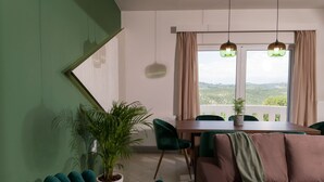 A modern living room with a dining area, green and brown furniture, a large window with a view, and hanging pendant lights. - Panoramic Sea Views, Pool, Garden, BBQ, TableTennis, Playground, Hammock (Chania)