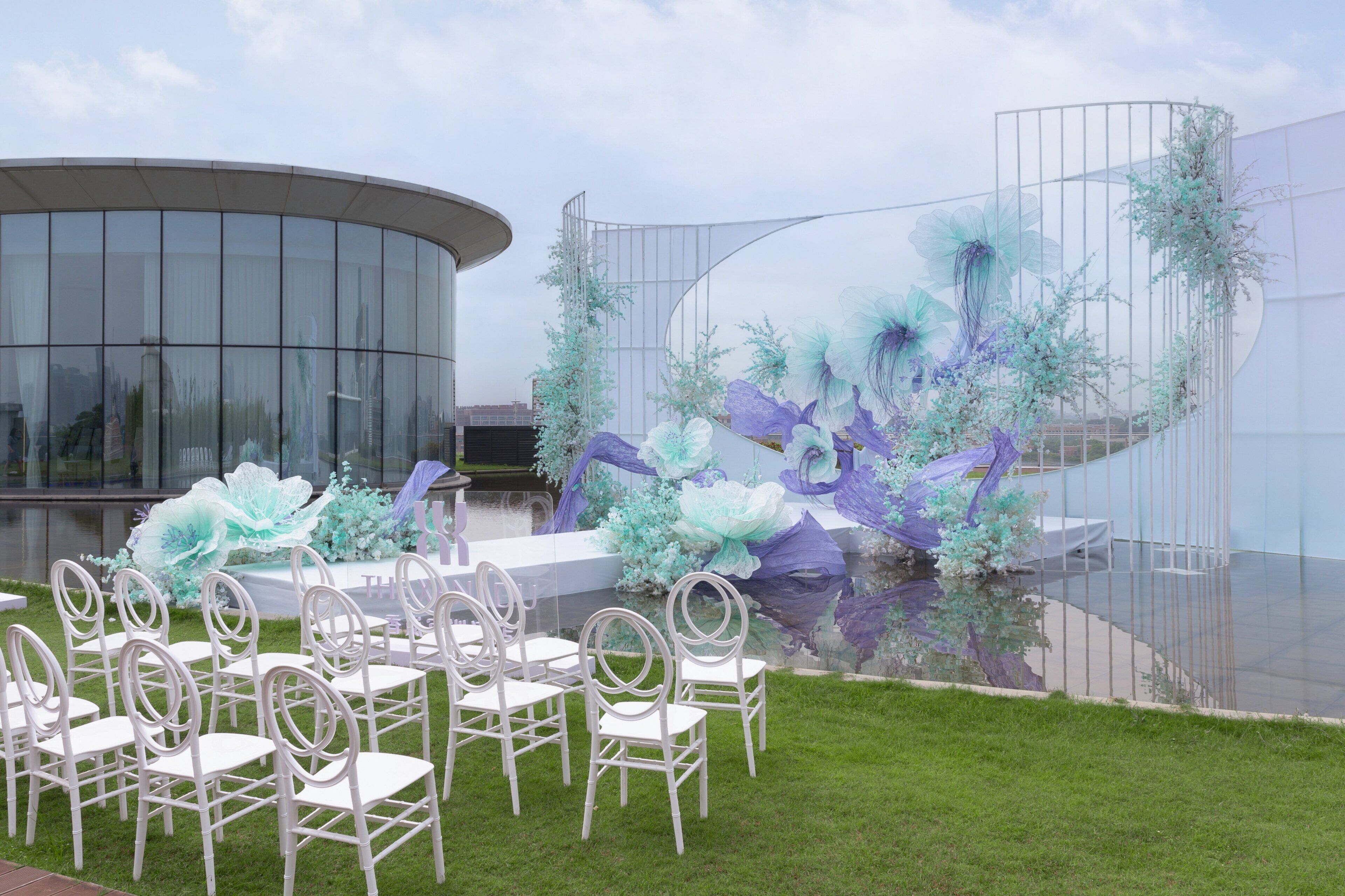 A modern outdoor venue with a circular glass building, white chairs arranged in rows, and decorative floral arrangements.