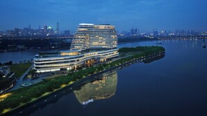 A modern, multi-story hotel with a unique curved architecture, situated by a river with a city skyline in the background. - Guangzhou Xanadu Hotel, Vignette Collection by IHG (Guangzhou)