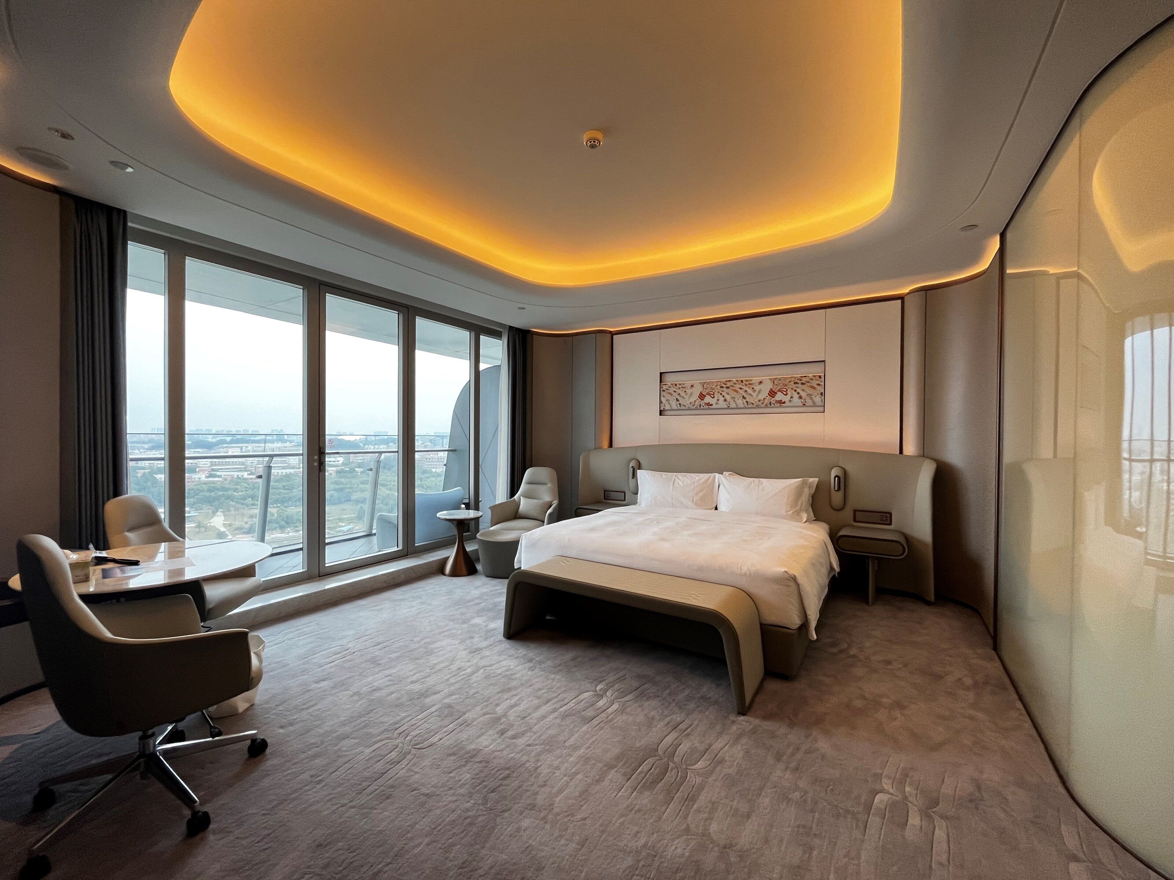 A modern hotel room with a large bed, a desk, a chair, and a view of the city.