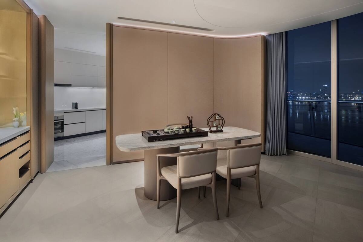 A modern dining area with a marble table and chairs, a view of the city at night, and a kitchen in the background.