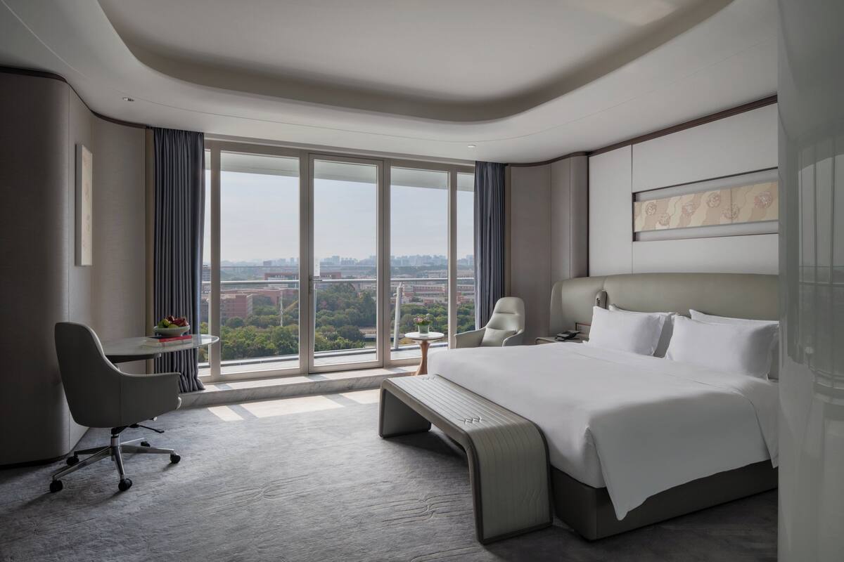 A modern hotel room with a large bed, a desk, a chair, and a view of the city.