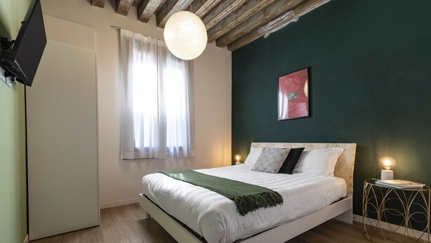 Cama bem arrumada com roupa de cama branca e manta verde, mesa de cabeceira com abajur, e janela com cortinas leves.