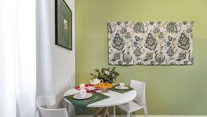 Uma sala de jantar com uma mesa redonda, cadeiras brancas, toalha de mesa verde e um painel floral na parede.