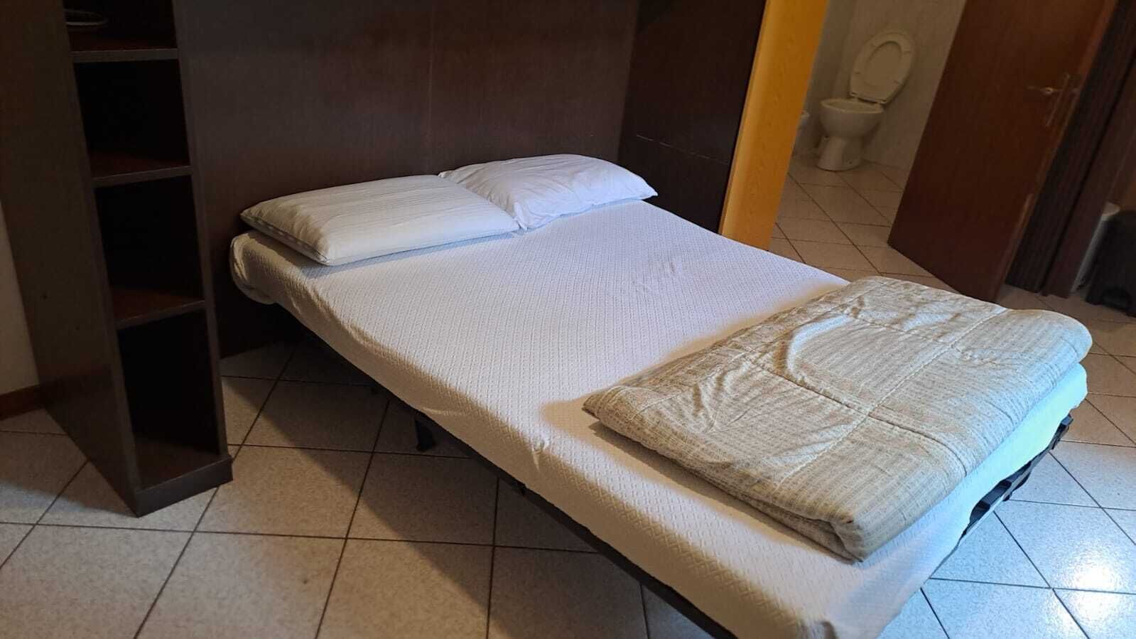 A single bed with a white mattress and pillows, a wooden headboard, a wooden wardrobe, and a tiled floor.