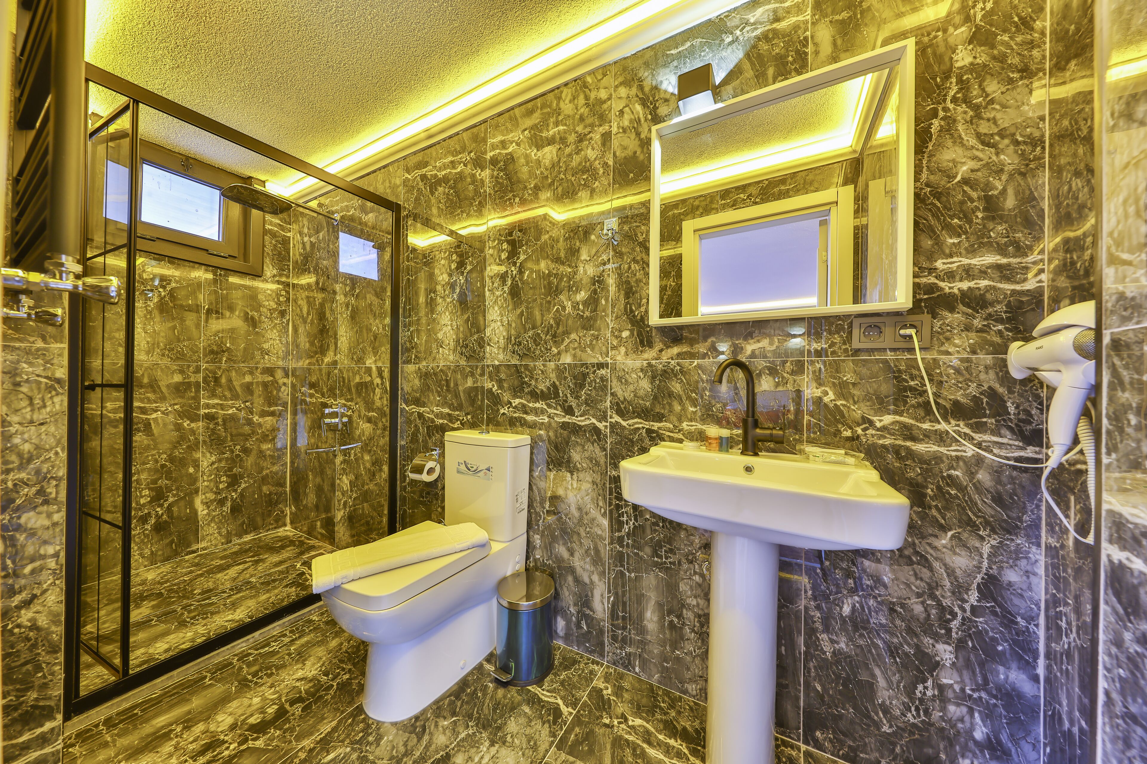 Baño con inodoro, lavabo y zona de ducha, con paredes de mármol y un espejo sobre el lavabo.