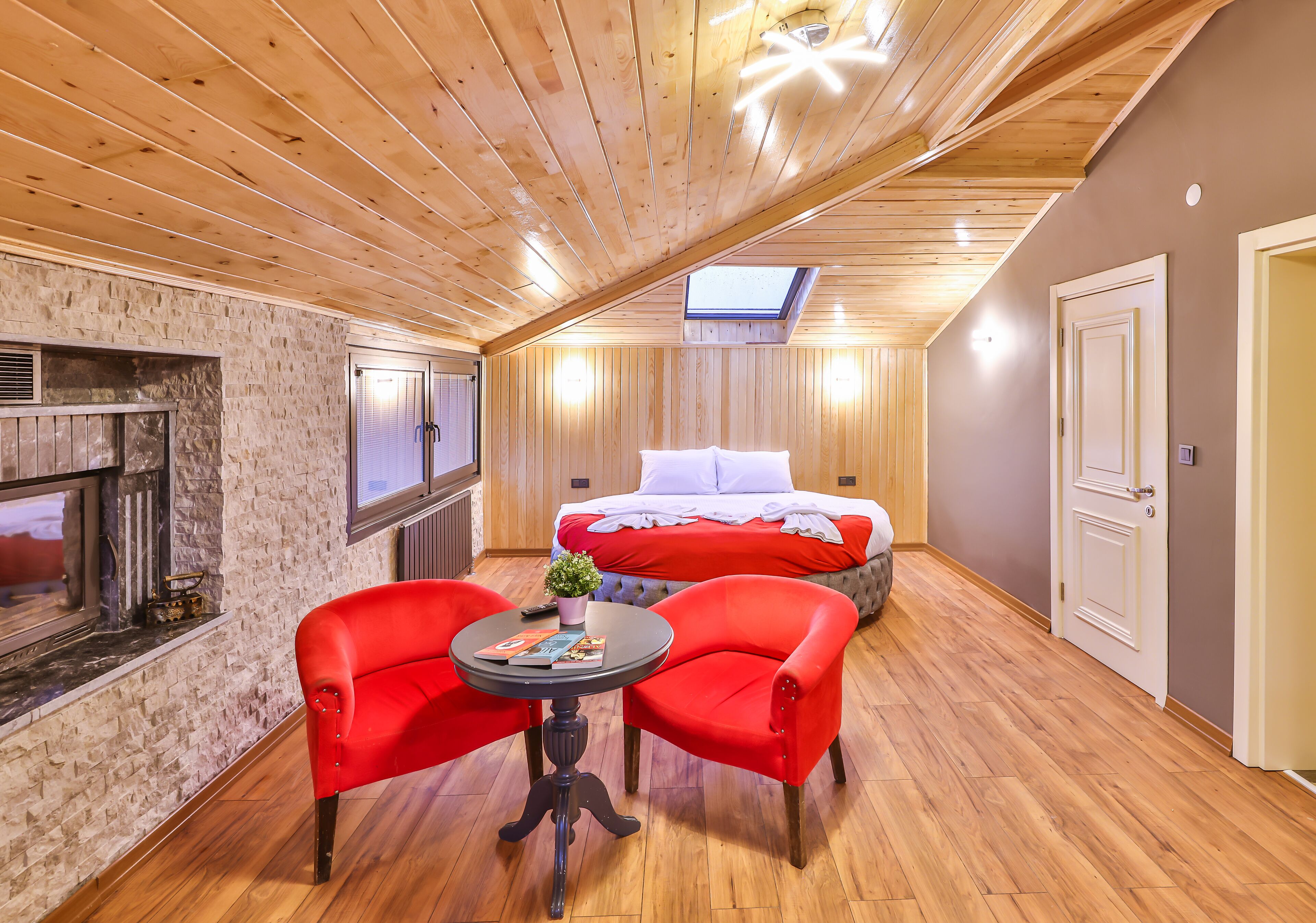 Una habitación acogedora con techo inclinado, una chimenea, una cama con ropa de cama roja y dos sillones rojos alrededor de una mesita.