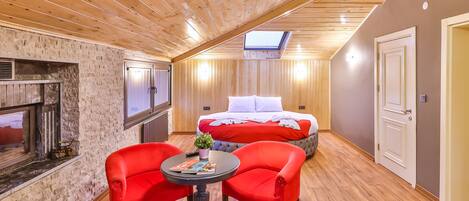Una habitación acogedora con techo inclinado, una chimenea, una cama con ropa de cama roja y dos sillones rojos alrededor de una mesita.