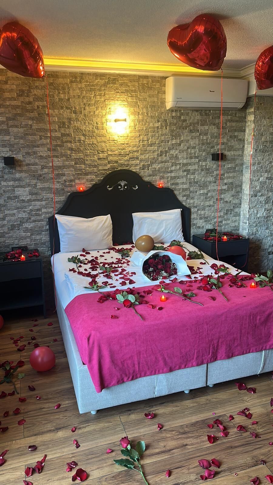 A bed with rose petals, a bouquet, and heart-shaped balloons in a room with a brick wall and an air conditioning unit.