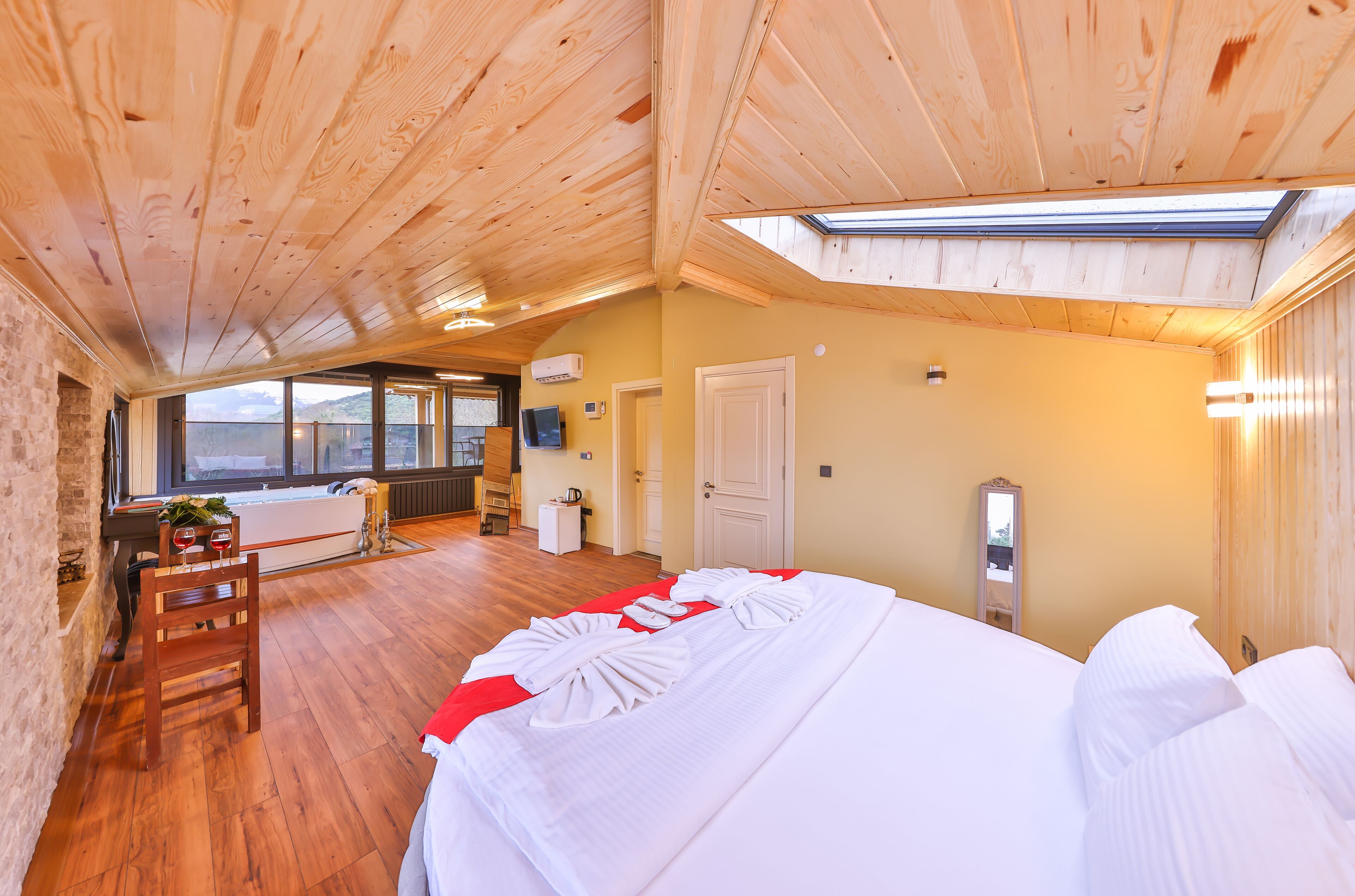 Techo de madera con tragaluces, cama con sábanas blancas, una silla de madera, una mesita con copas de vino y vistas al exterior.