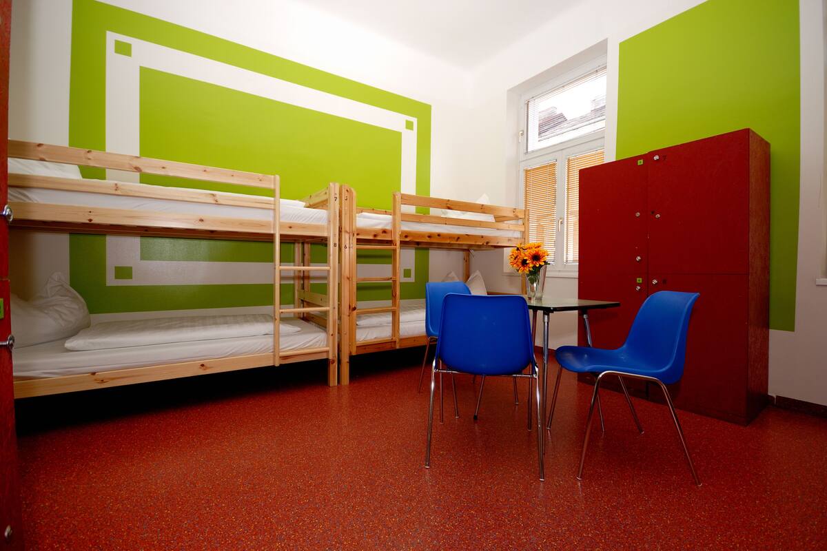 A bunk bed room with a table and chairs, a red cabinet, and a window with blinds.