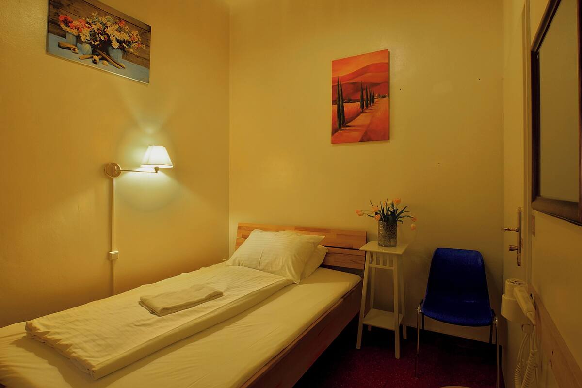 A small, neatly made bed with a white comforter and pillows, a bedside lamp, a small table with a vase of flowers, and a blue chair.