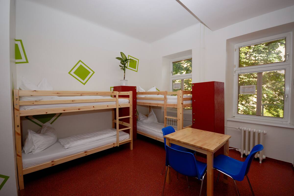 A dormitory room with bunk beds, a small table, chairs, a radiator, and a window with blinds.