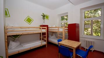 Un dormitorio con literas, una mesita, sillas, un radiador y una ventana con persianas.