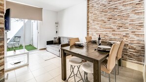 Una zona de menjador moderna amb una taula de fusta i quatre cadires, un televisor muntat sobre una paret blanca i una paret a la dreta amb un disseny de rajoles.