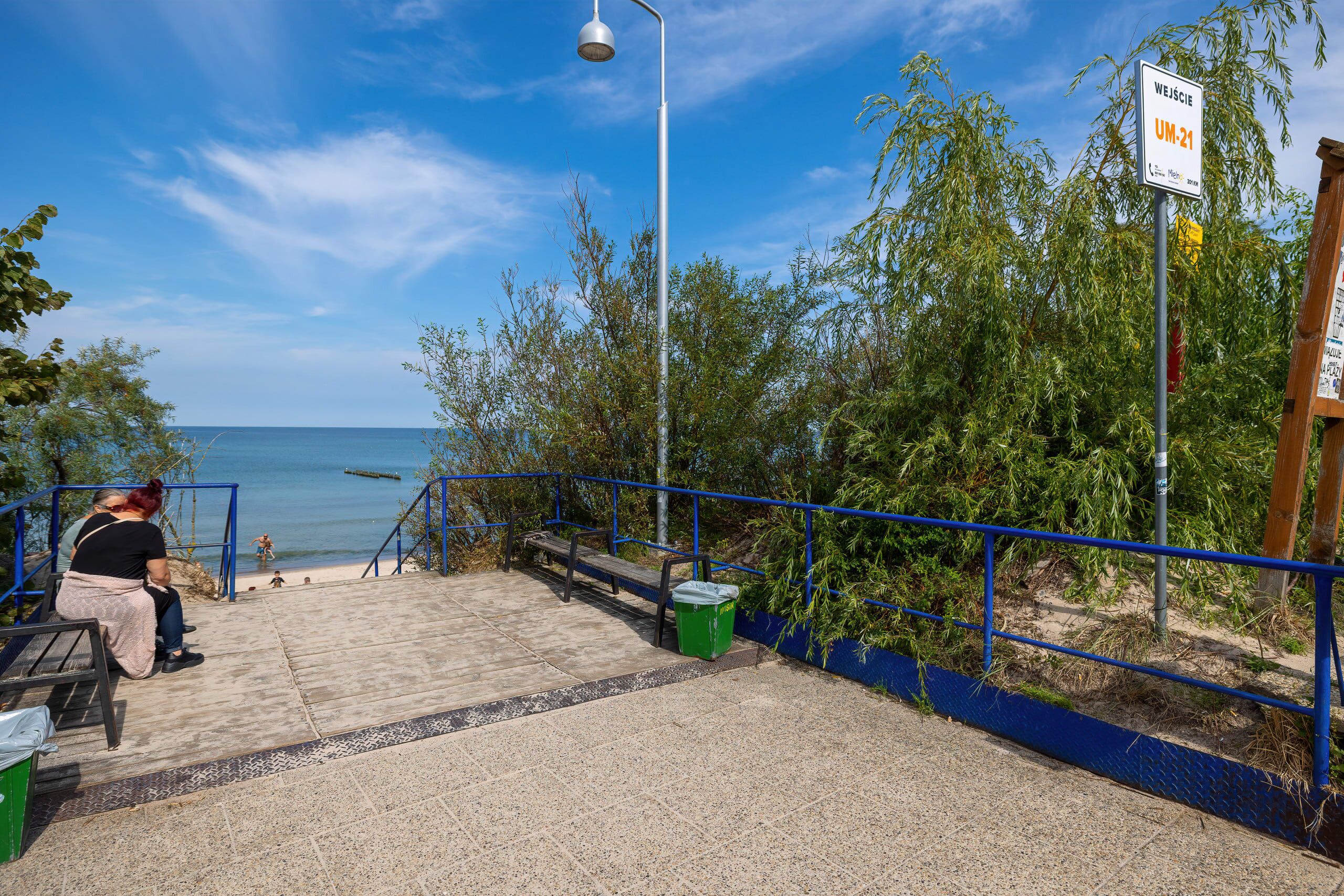 A beachside walkway with a metal lamp post, a trash bin, and a bench.