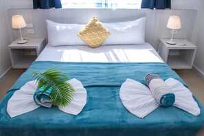 Una cama bien hecha con una colcha color esmeralda, almohadas blancas y una almohada decorativa. Hay dos mesitas de noche con lámparas, una planta de palma y toallas enrolladas sobre la cama.