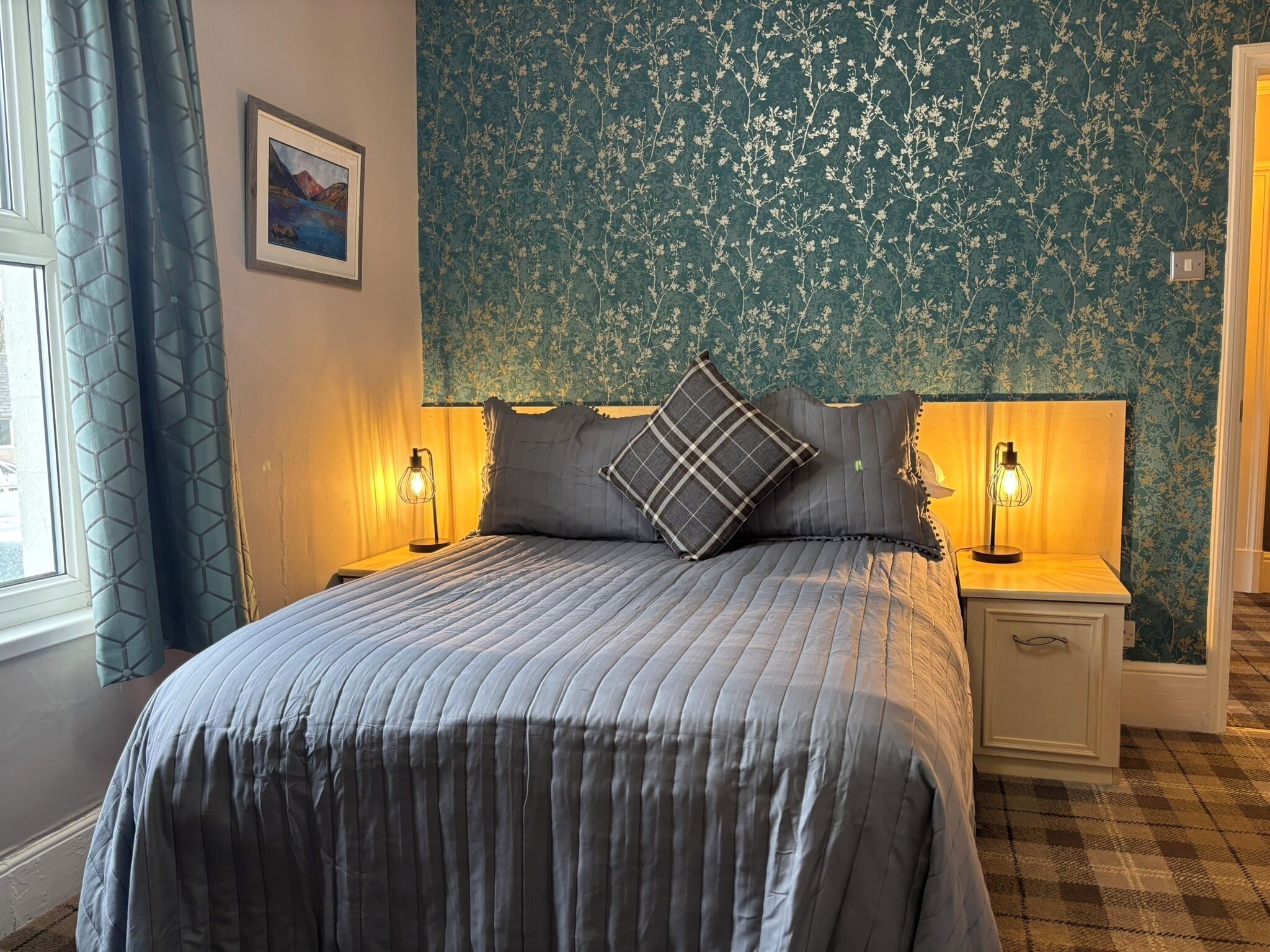 A bedroom with a bed, two bedside lamps, a headboard, a framed picture, and a window with curtains.