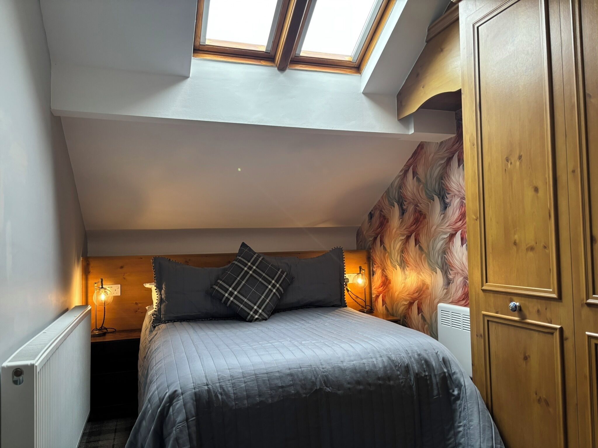 A bedroom with a skylight, a bed with a quilted cover, a radiator, and a wall with a feather-like pattern.