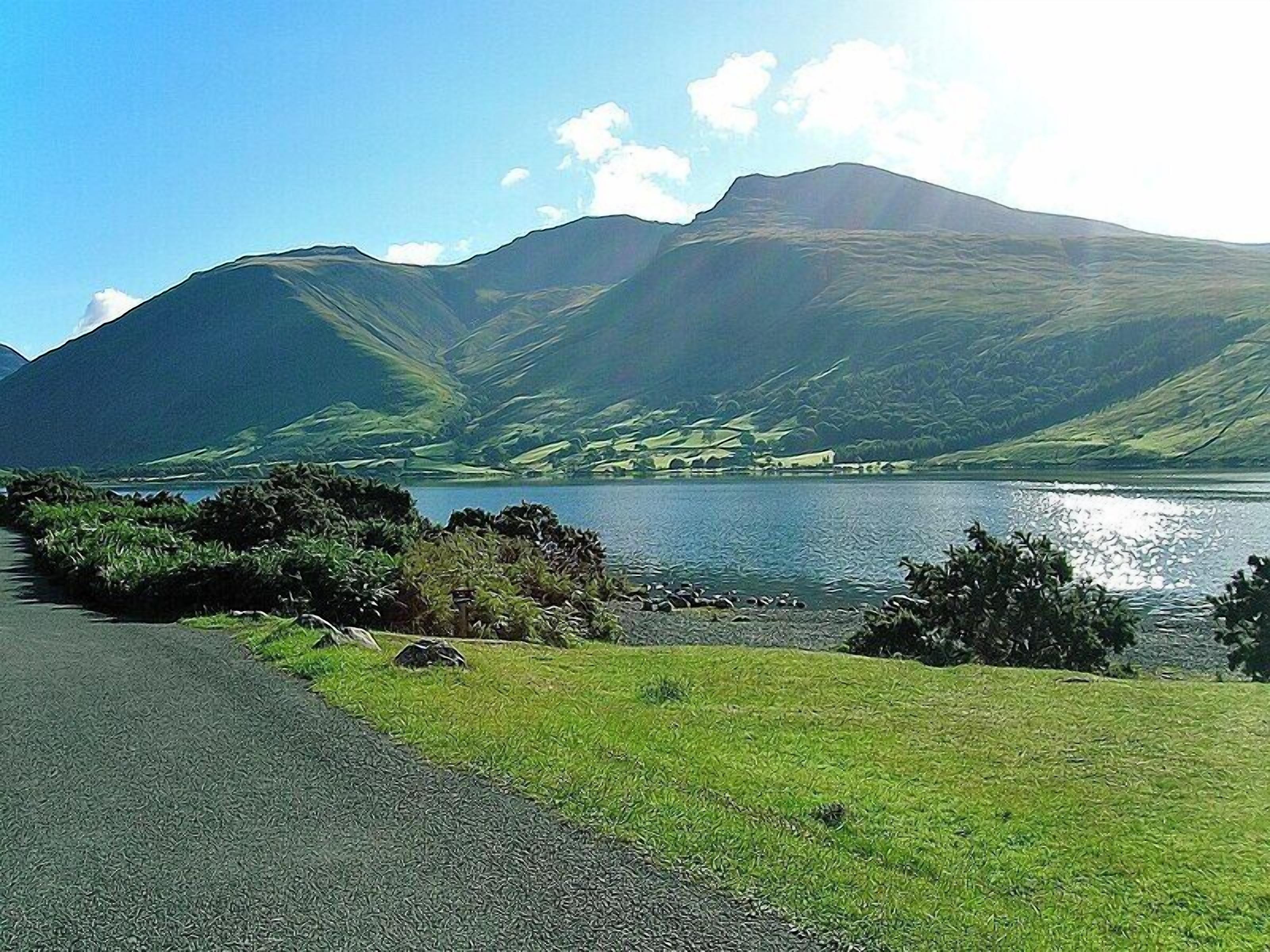 A scenic view of a lake with mountains in the background, a grassy area in the foreground, and a road leading towards the lake.