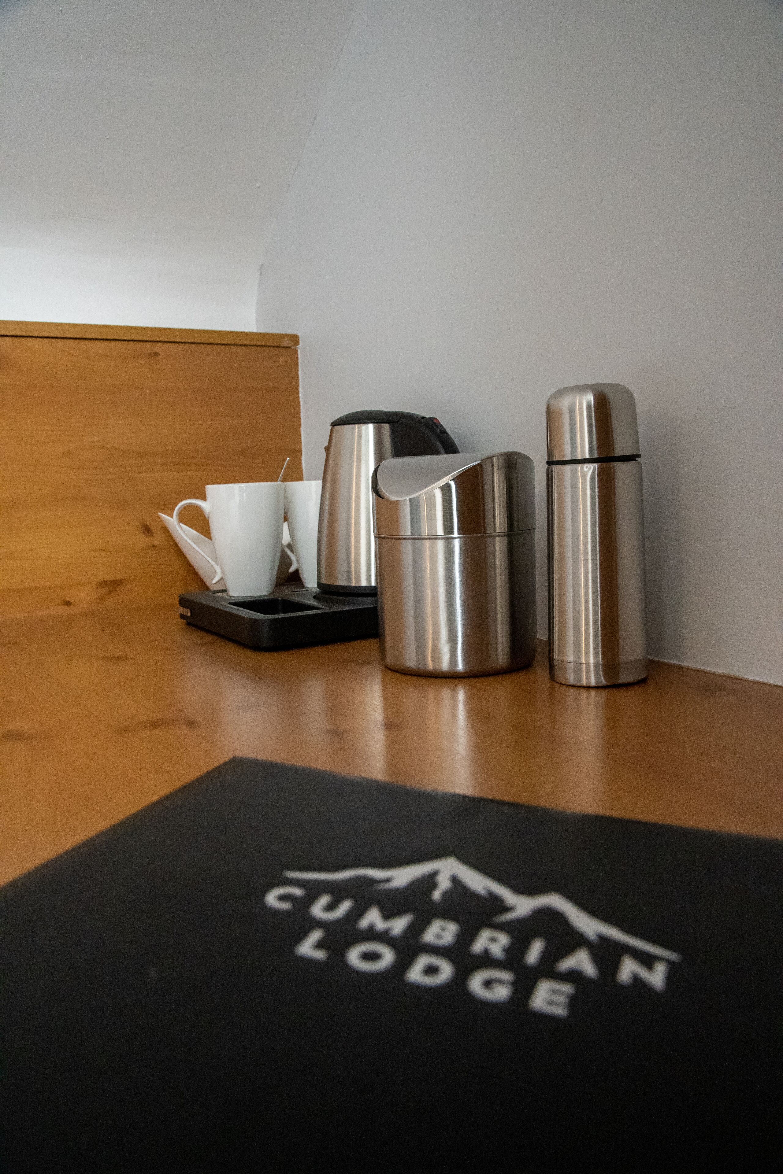A wooden table with a black placemat featuring the logo "CUMBRIAN LODGE". On the table, there are two stainless steel thermoses, a black electric kettle, and two white cups with a spoon.