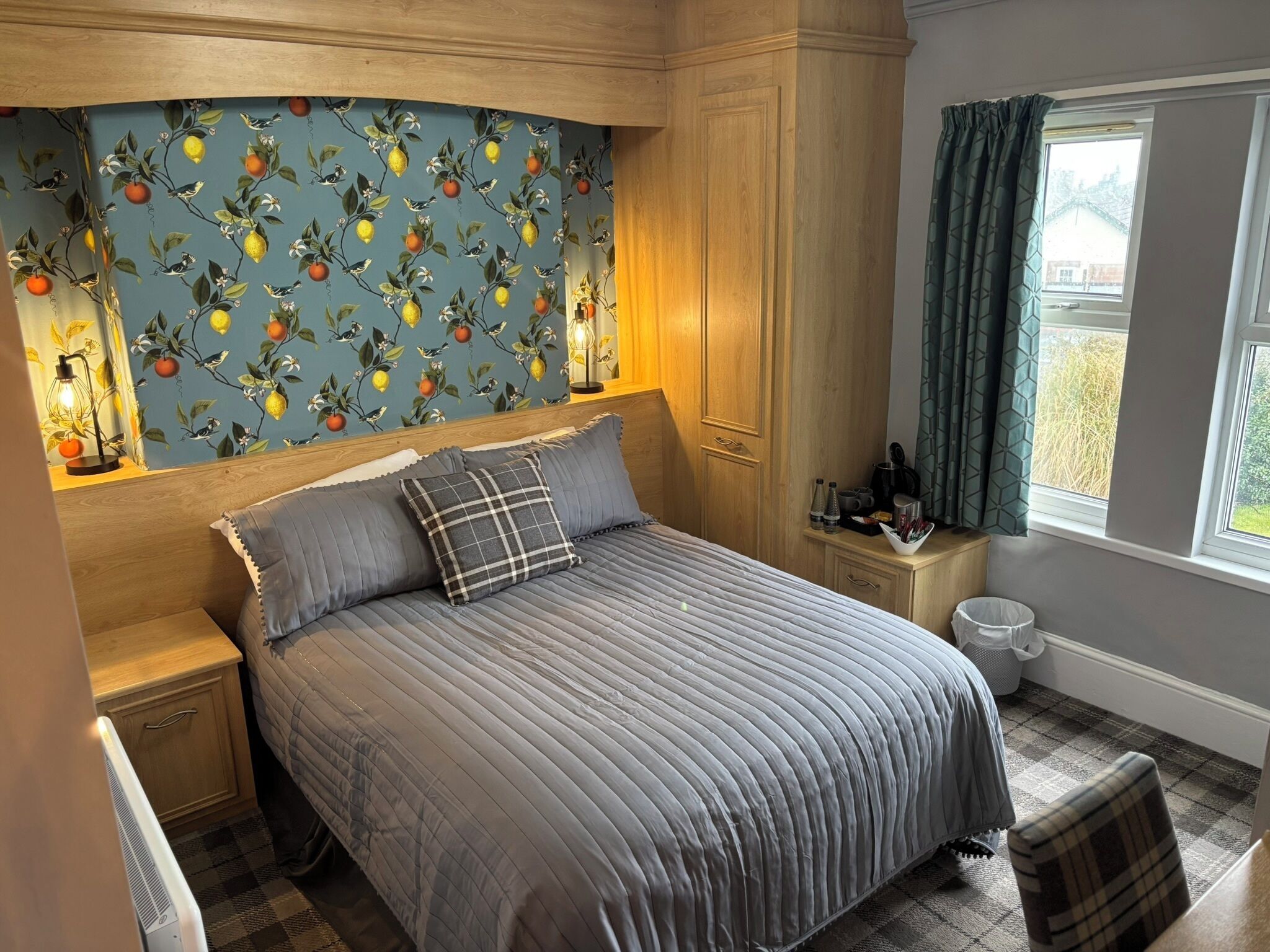 A bedroom with a bed, a nightstand, a dresser, a window with curtains, and a wall with a fruit and bird pattern.