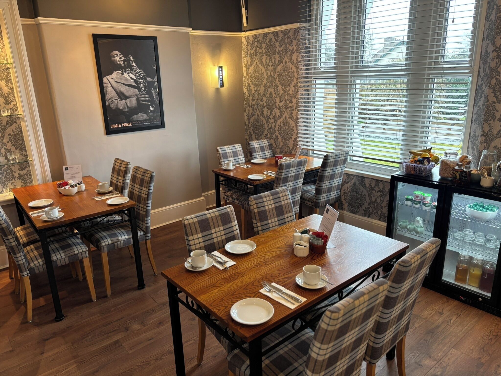 A dining area with wooden tables and plaid chairs, a poster on the wall, and a refrigerator with beverages.