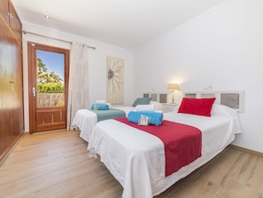 A hotel room with a wooden door, two beds with white bedding, a red throw, and a view of palm trees. - Villa Can Segue · Villa with private pool in Alcúdia (Alcúdia)