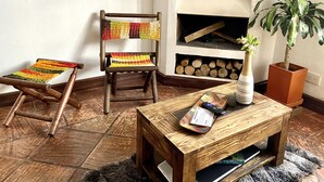 A wooden chair with a colorful woven seat and backrest, a small wooden table with a remote control and a card, and a potted plant beside a fireplace.