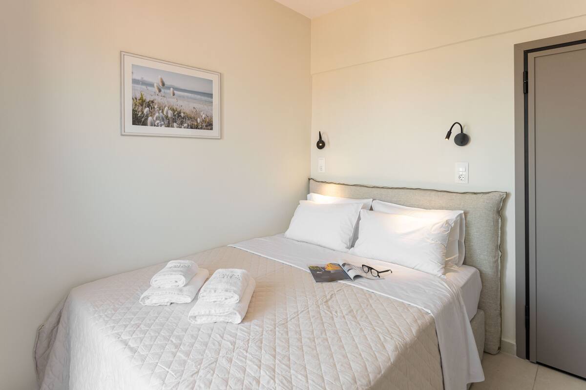 Una cama bien hecha con sábanas blancas, un cuadro en la pared y unos auriculares en la mesita de noche.