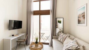 A modern living room with a white sofa, a TV on a white console, a wicker side table with a plant, and a view of the outdoors through large windows. - Palm Residences (Kassandra)