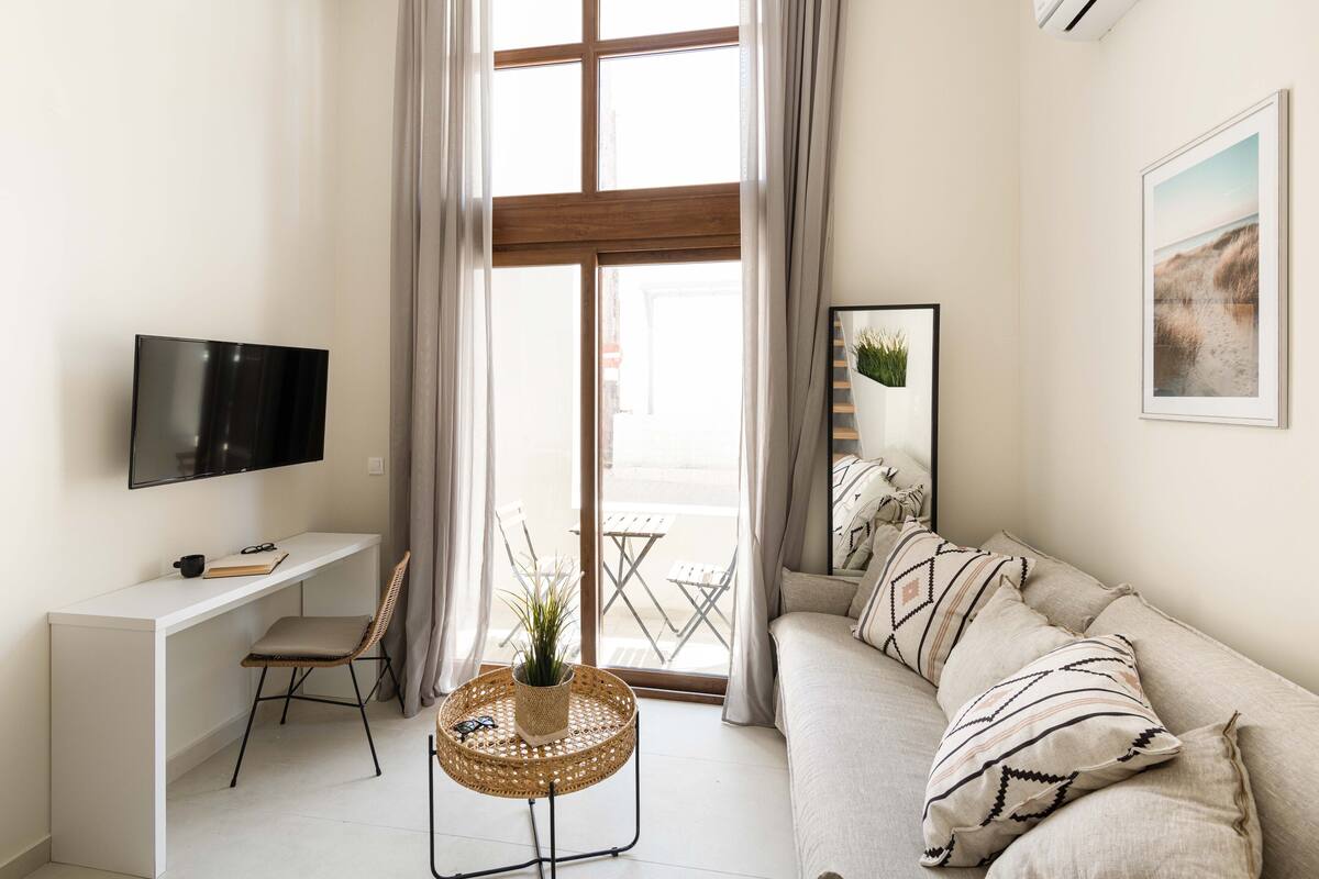 Una sala de estar moderna con un sofá blanco, un televisor sobre un mueble blanco, una mesita de mimbre con una planta y vistas al exterior a través de ventanas amplias.