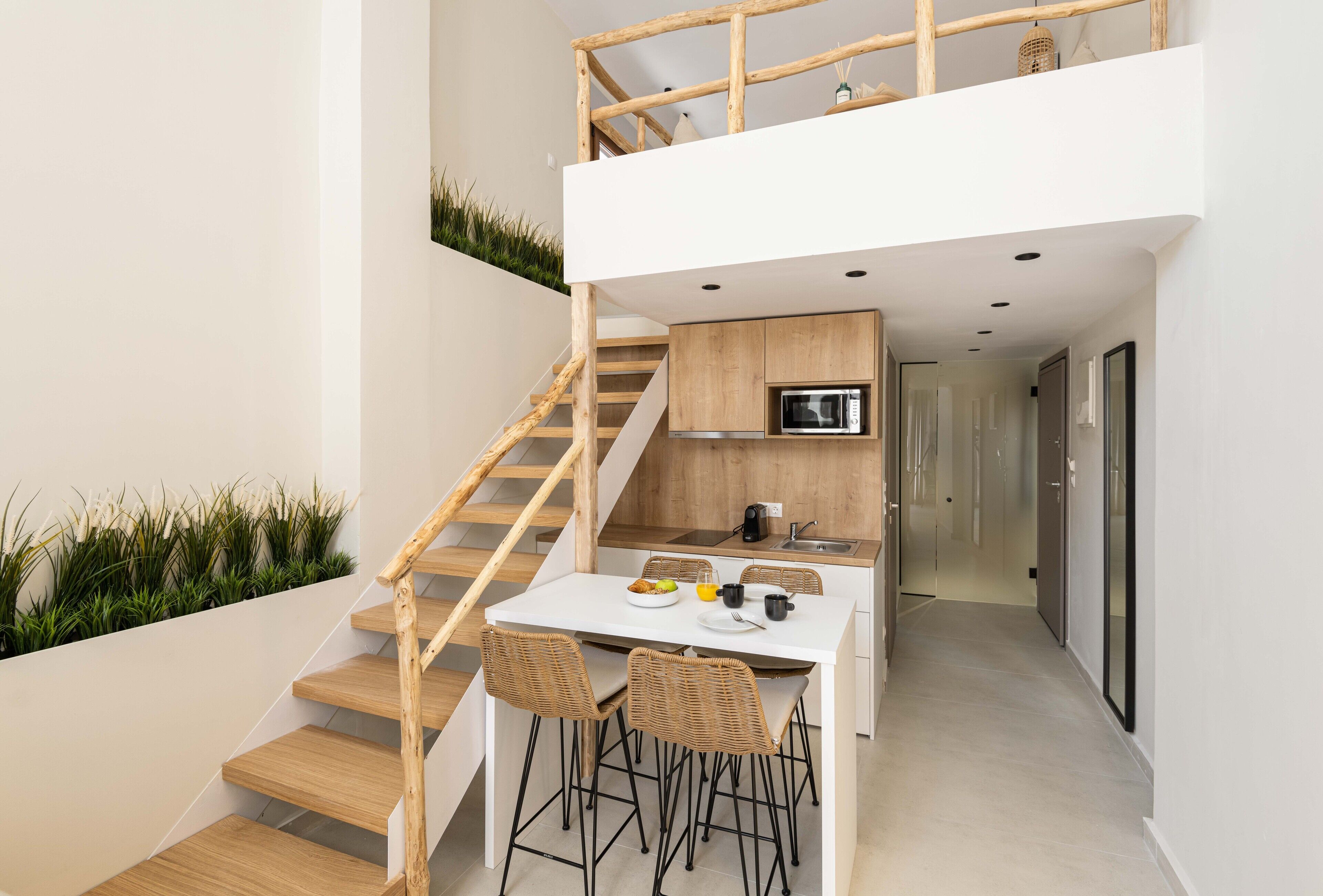 A modern kitchen with a white countertop, wooden cabinets, and a built-in oven. There is a dining area with a white table and four wicker chairs. A wooden staircase leads to an upper level.