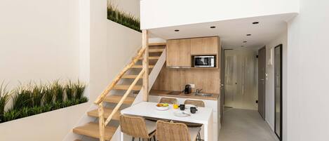 A modern kitchen with a white countertop, wooden cabinets, and a built-in oven. There is a dining area with a white table and four wicker chairs. A wooden staircase leads to an upper level.