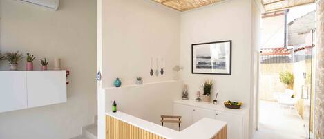 A modern reception area with a wooden reception desk, white walls, and a ceiling with a natural fiber texture. There are potted plants on a shelf and a framed picture on the wall.