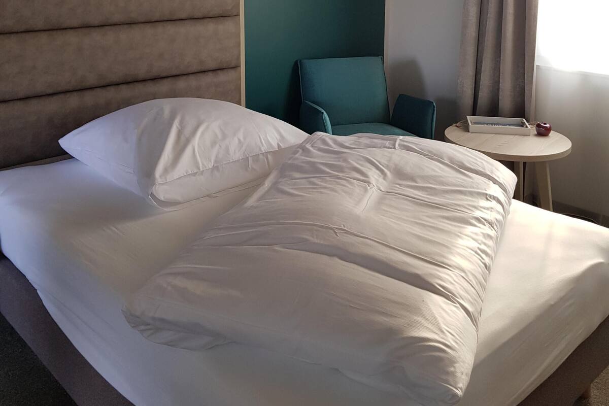 A hotel room with a bed, a chair, a small table, and a window with curtains.