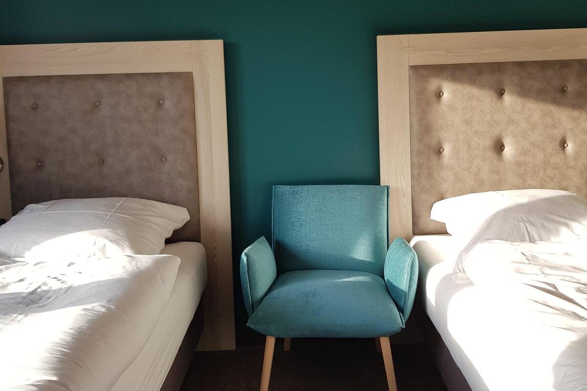 A room with two beds, a blue chair, and teal walls.