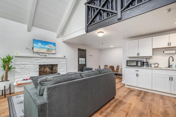A modern living room with a grey sectional sofa, a fireplace, and a kitchen area with white cabinets and a microwave.