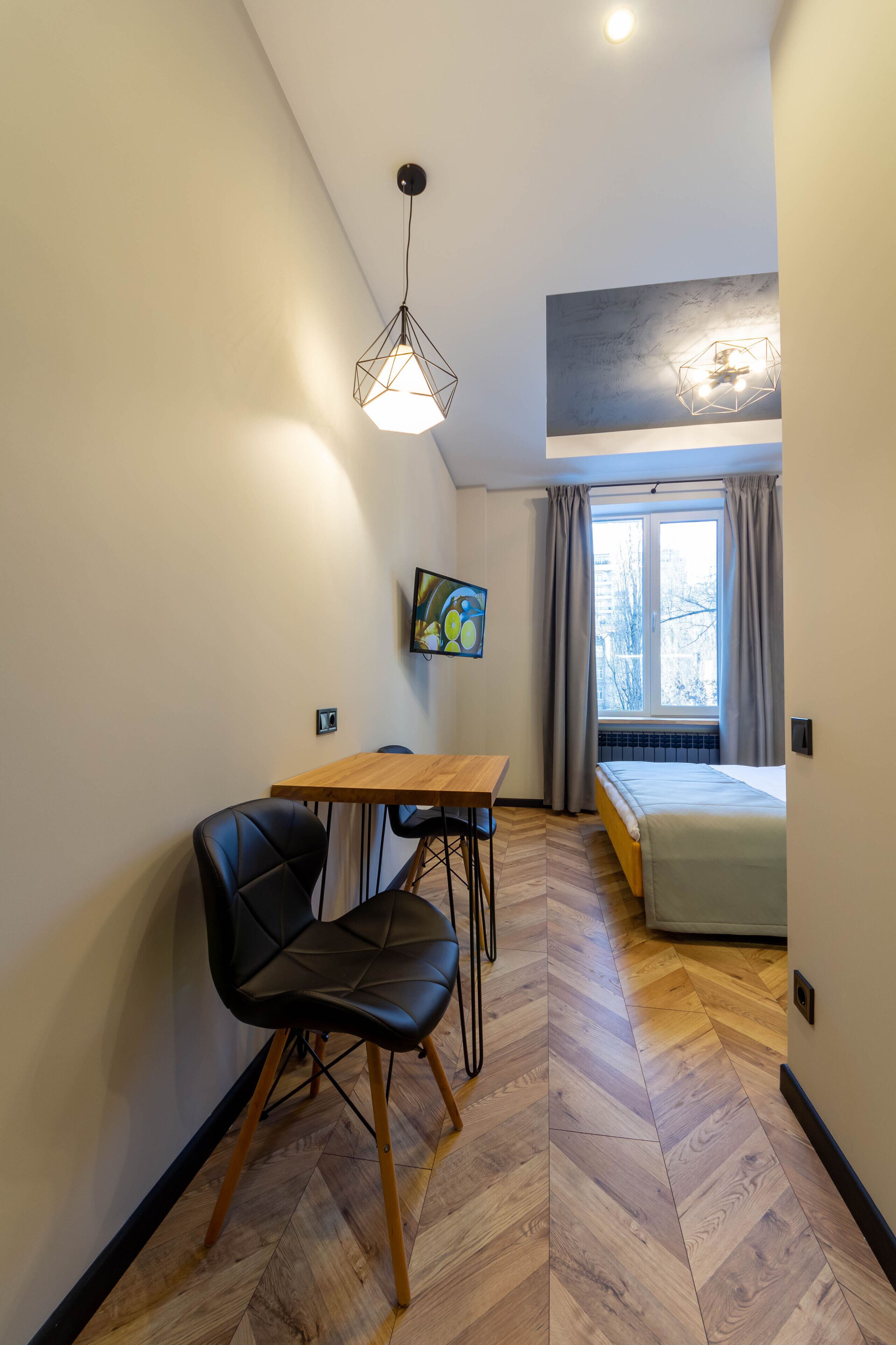 Una habitación pequeña y moderna con una mesa de madera y una silla negra, una cama con cabecera gris, un cuadro en la pared y una ventana con cortinas.