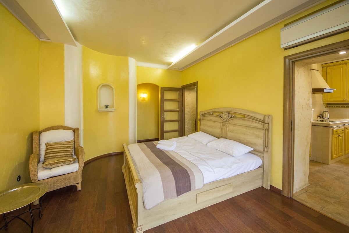 A bedroom with a bed, a chair, a small table, and a kitchen area visible through an open door.