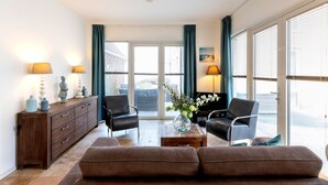 A living room with a brown sofa, two armchairs, a wooden coffee table, a cabinet with decorative items, and a view through large glass doors. - Flat Fish (Egmond aan Zee)